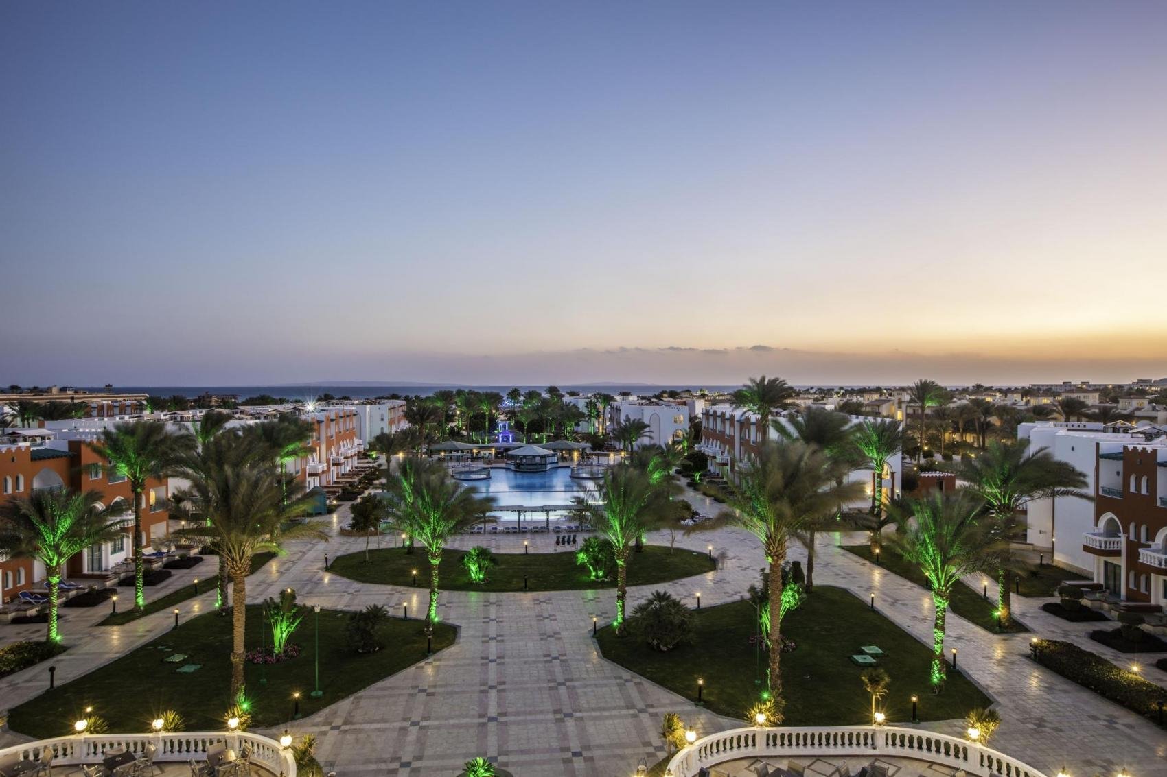 Vakarā skats uz viesnīcu kompleksu ar palmām un apgaismotu laukumu pie ūdens. Fons veido krāšņa saulrieta ainava ar jūras horizontu. panoramic view with beach and pools