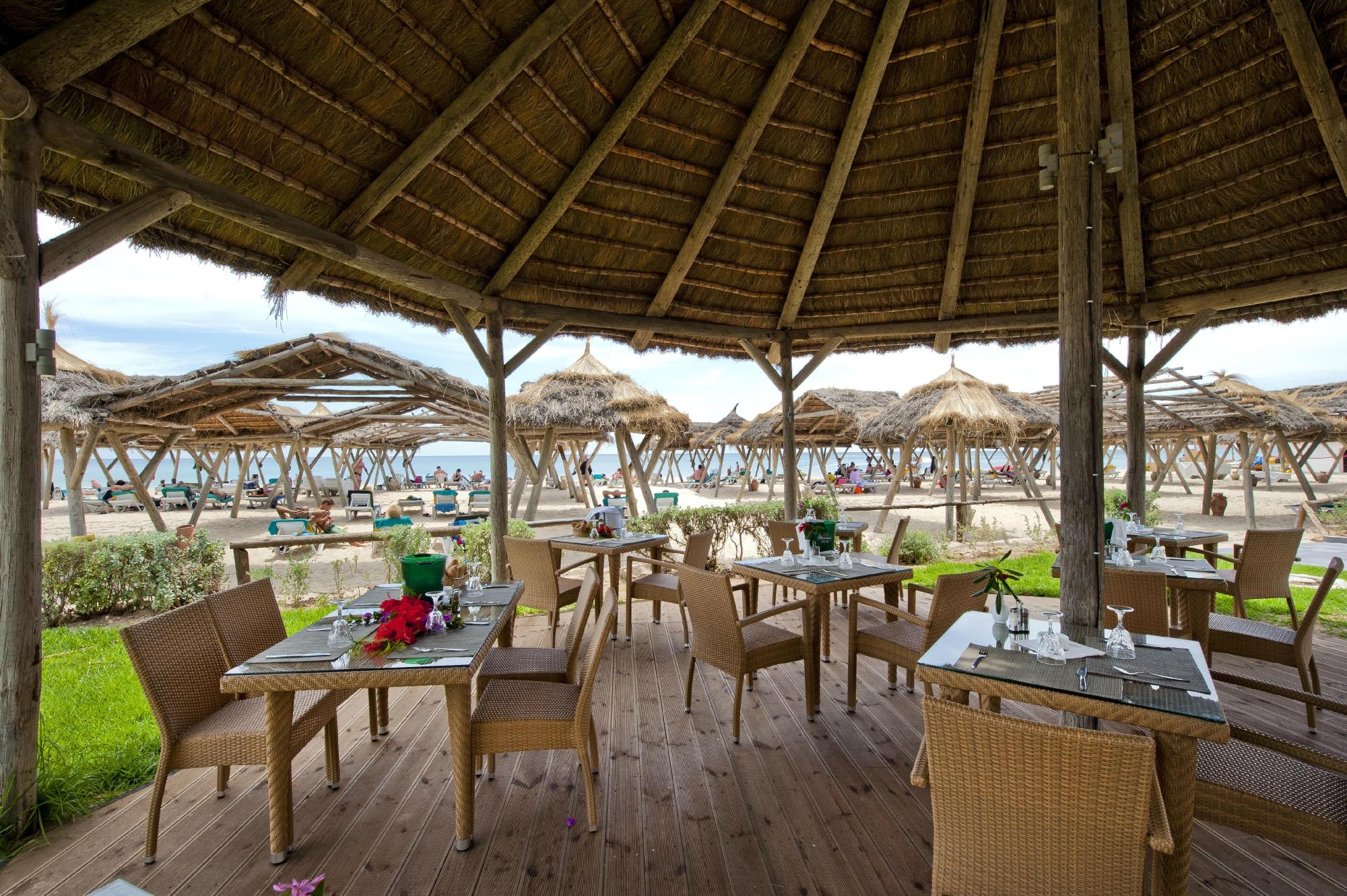 Vasaras restorāns ar koka terasi, no kuras paveras skats uz pludmali un atpūtniekiem. Galdi ir klāti ar ziediem un traukiem, radot relaksējošu atmosfēru. panoramic view with beach and pools
