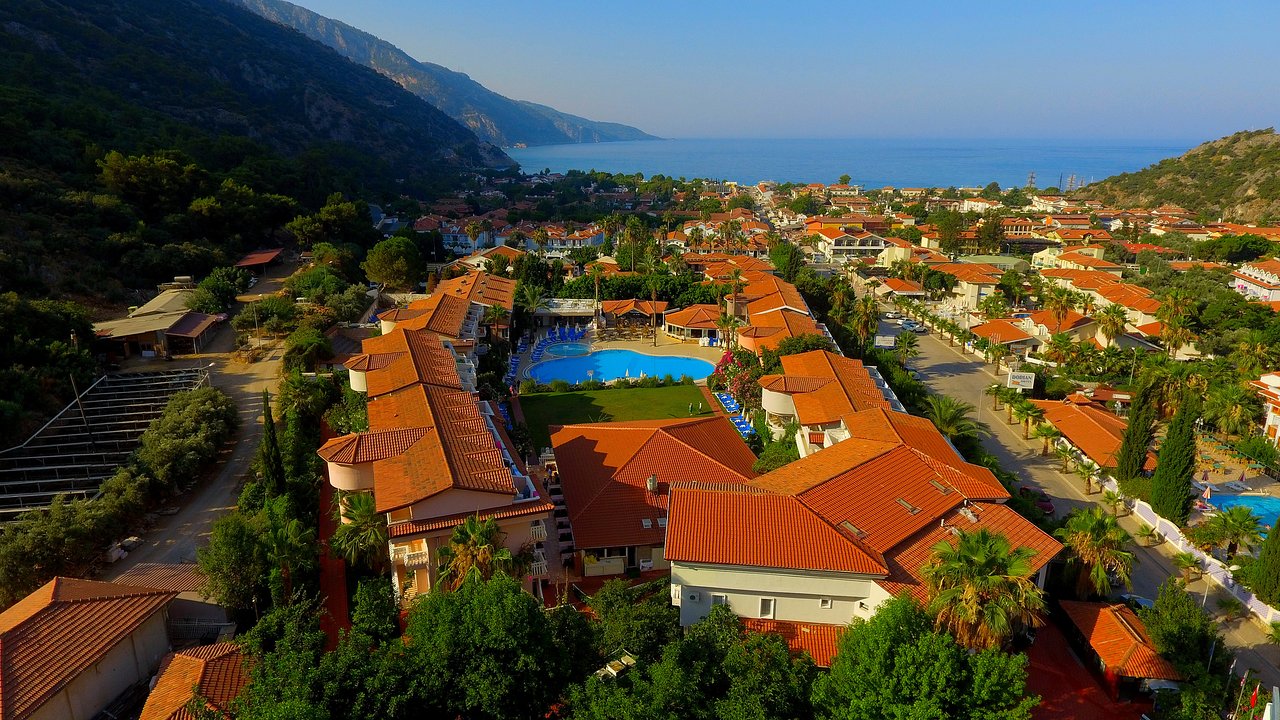 Ainava ar viesnīcu kompleksu pie jūras, kurā redzami sarkanie jumti un peldbaseins, apkārt augstie kalni un zāles. panoramic view with beach and pools