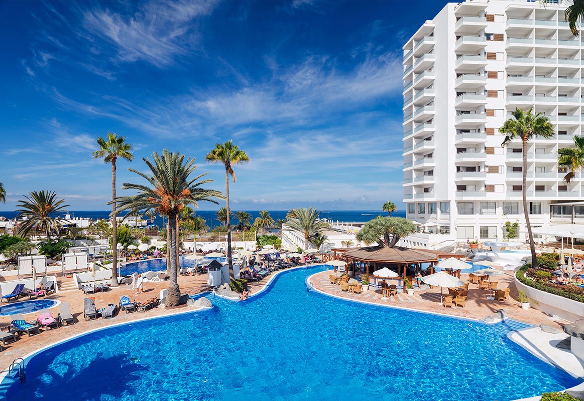 Viesnīcas komplekss ar peldbaseinu, palmām un saulainu debesu fonu, kas atrodas pie pludmales. Skats uz jūru un atpūtas zonas. panoramic view with beach and pools